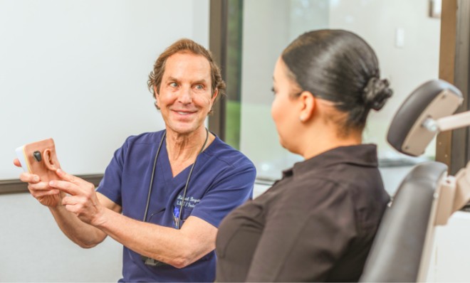 A doctor in navy scrubs, wearing blue gloves, holds a medical tool, attentively speaking to a seated patient in an office. Certificates are on the wall.